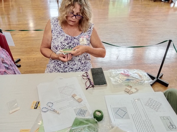 A member of the craft club examining her Hardanger embroidery closely, with printed charts and green Pearl cotton thread on the table