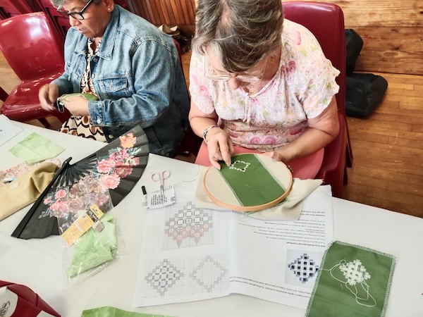 A stitcher working kloster blocks in green fabric using an embroidery hoop, with Hardanger charts spread beside her