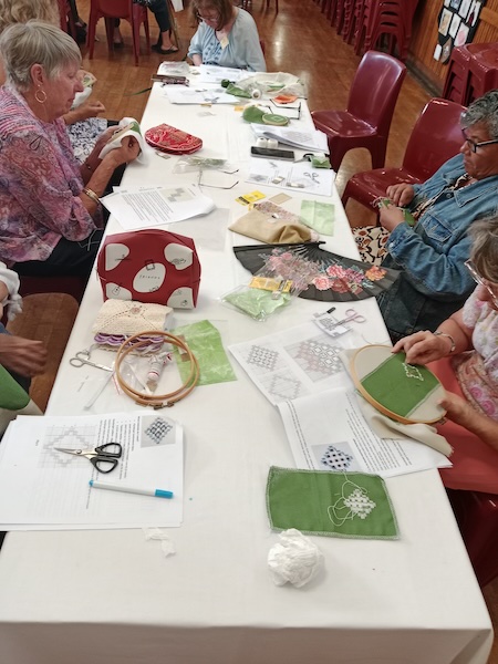 Members of the South Peninsula Handcraft Centre in Fish Hoek, South Africa, learning Hardanger embroidery around a table with printed charts, green fabric, embroidery hoops and scissors