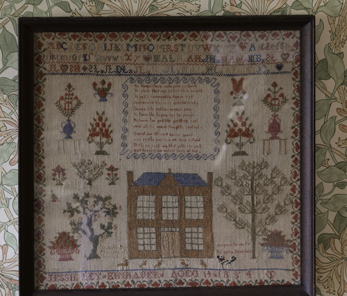Jessie's 1854 cross stitch sampler on linen with a spotty dog below the house